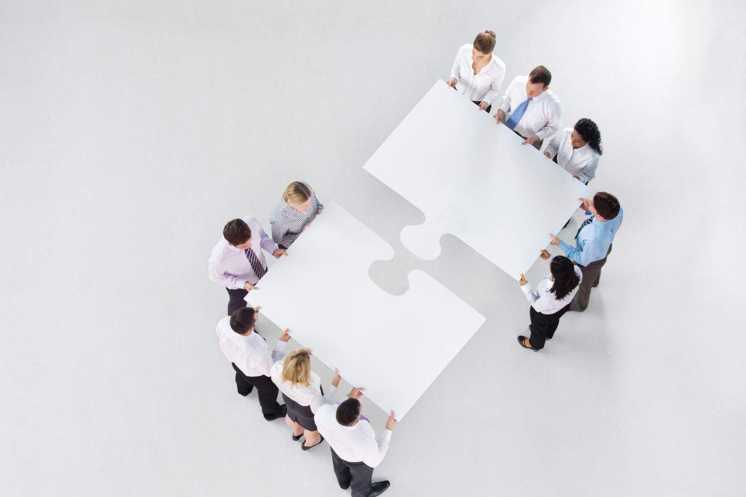 Overhead shot of two groups of businesspeople holding a large jigsaw piece coming together to merge denoting teamwork.