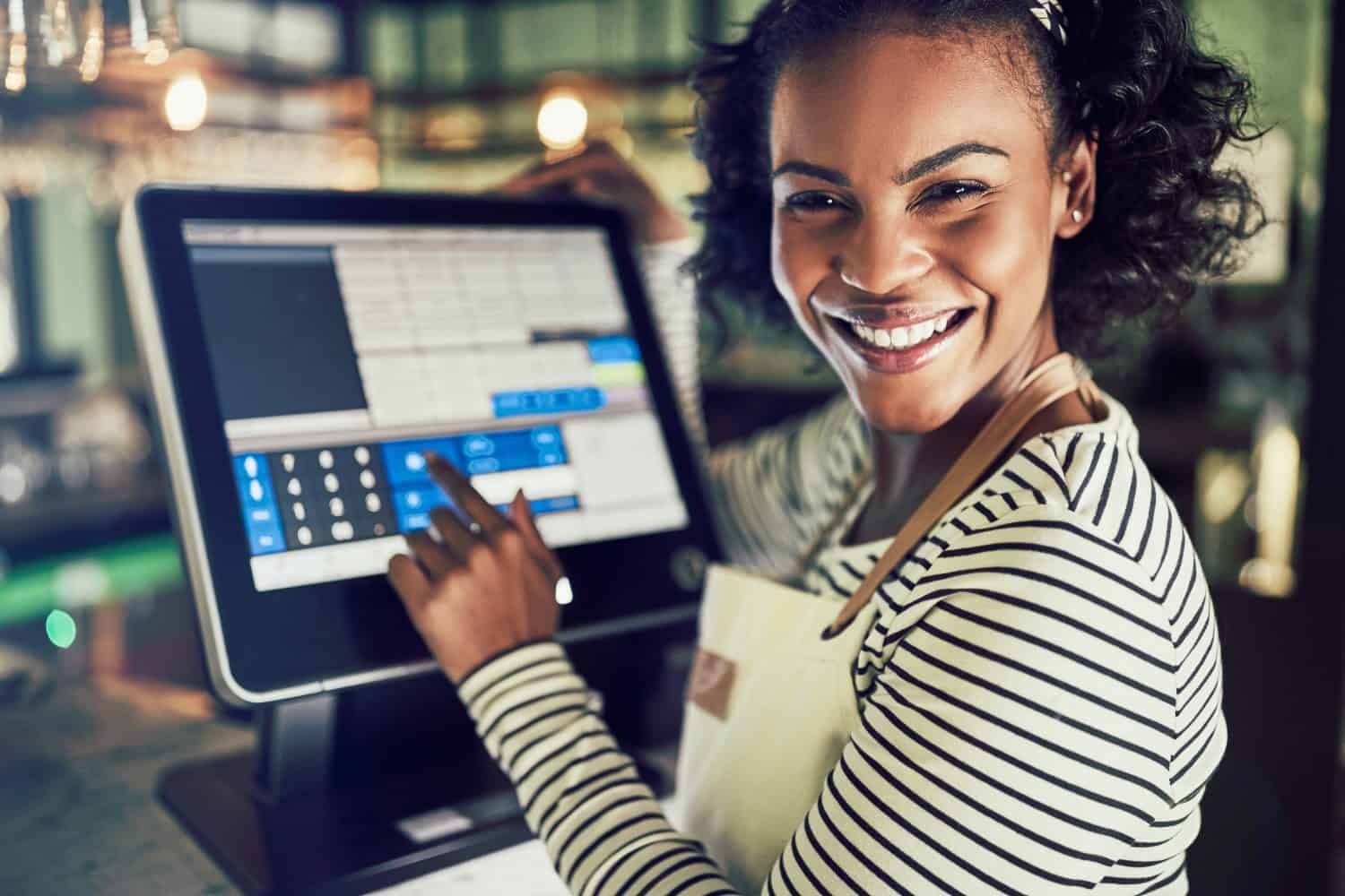 Smiling young African waitress wearing an apron working in a trendy restaurant and using a touchscreen point of sale terminal