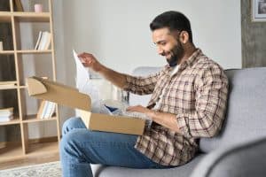 Happy smiling indian man opening box with ordered goods gifts, presents at home on couch. Online shopper male customer opening online shop parcel. International delivery service concept.