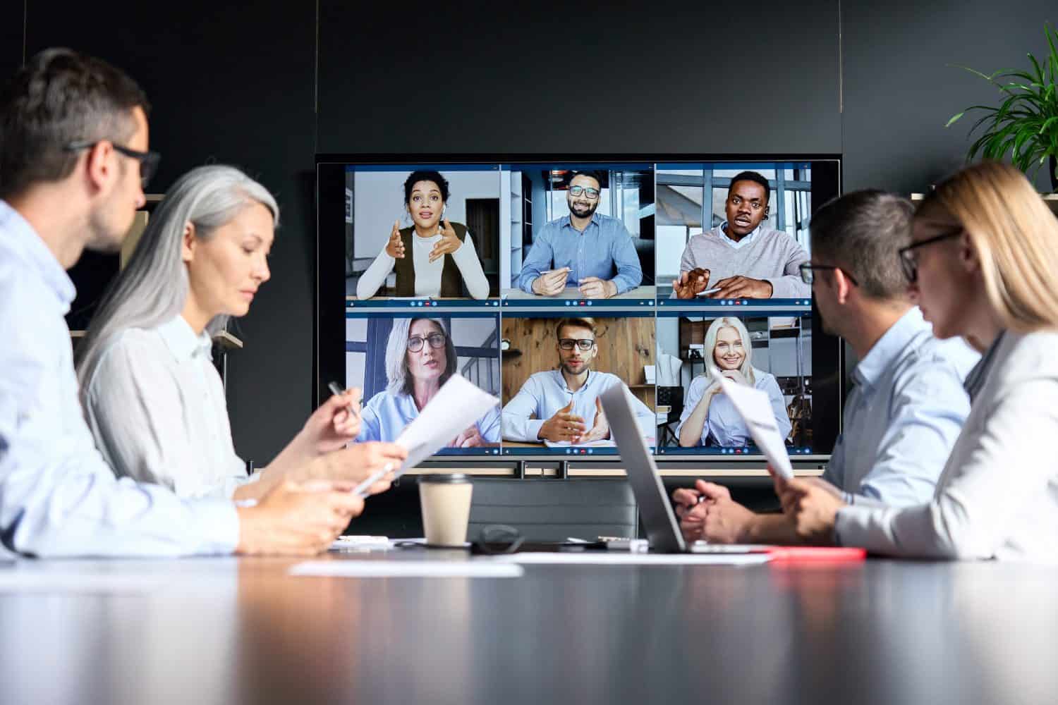 Global corporation online videoconference in meeting room with diverse people sitting in modern office and multicultural multiethnic colleagues on big screen monitor. Business technologies concept.