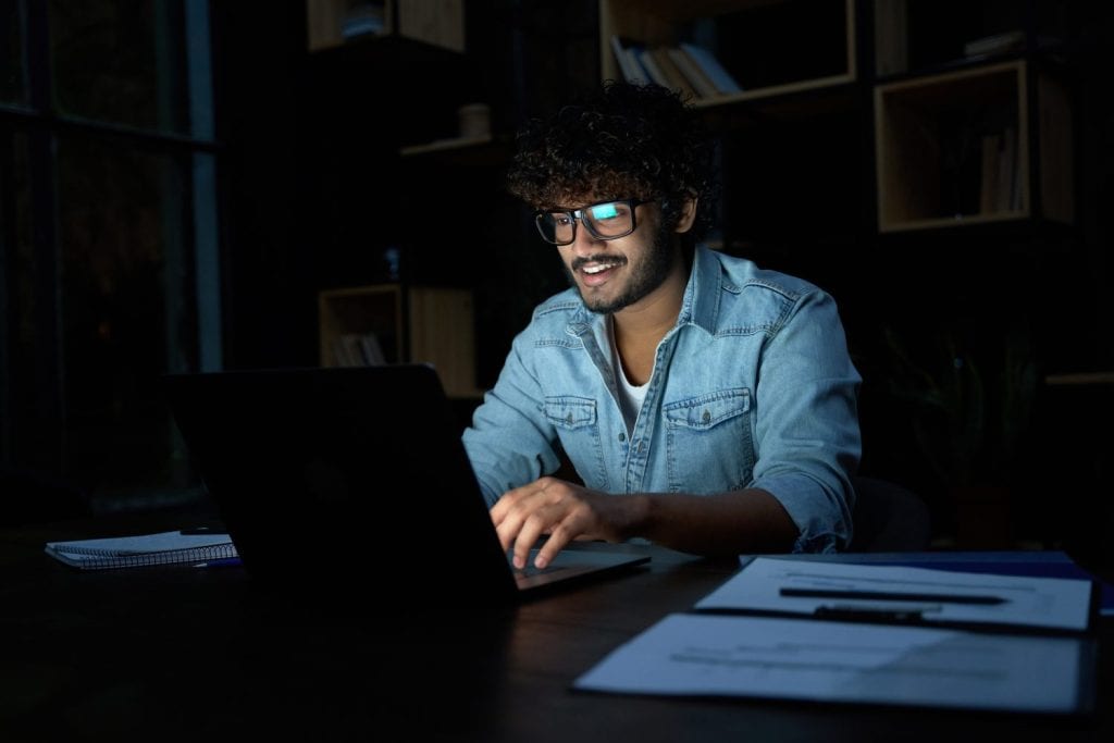 Smiling young curly indian latin ethnic business man or student wearing glasses remote working overtime, learning online late at night at home or in dark office using laptop computer at workplace.