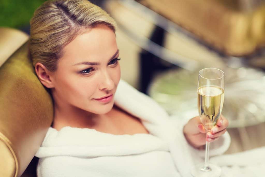 people, beauty, lifestyle, holidays and relaxation concept - beautiful young woman in white bath robe lying on chaise-longue and drinking champagne at spa