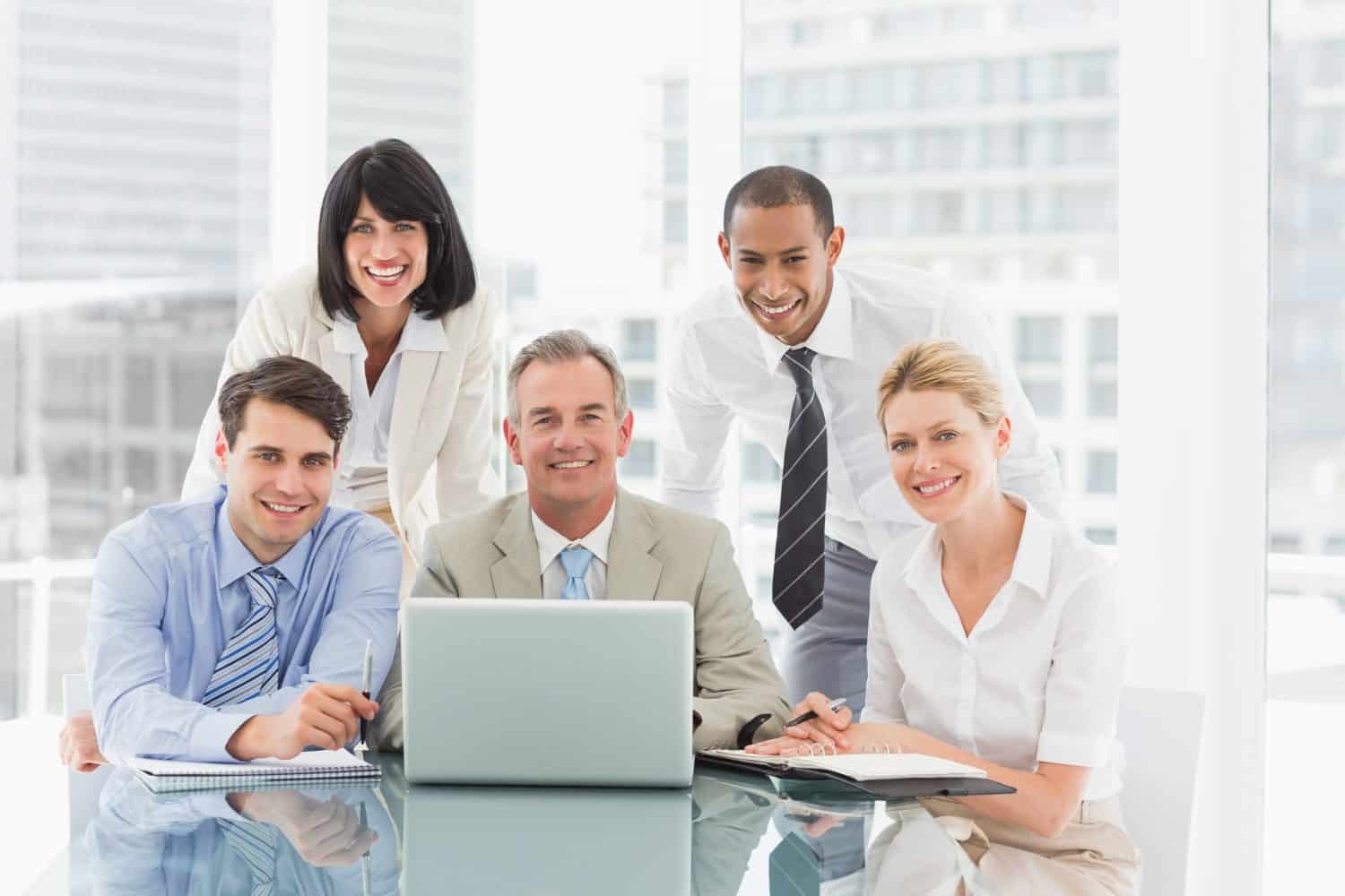 Happy business people gathered around laptop looking at camera in the office