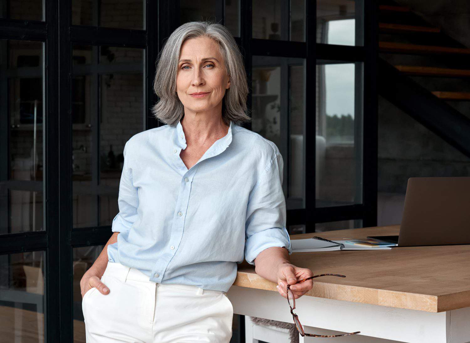 Confident stylish european mature middle aged woman standing at workplace. Stylish older senior businesswoman, 60s gray-haired lady executive leader manager looking at camera in office, portrait.