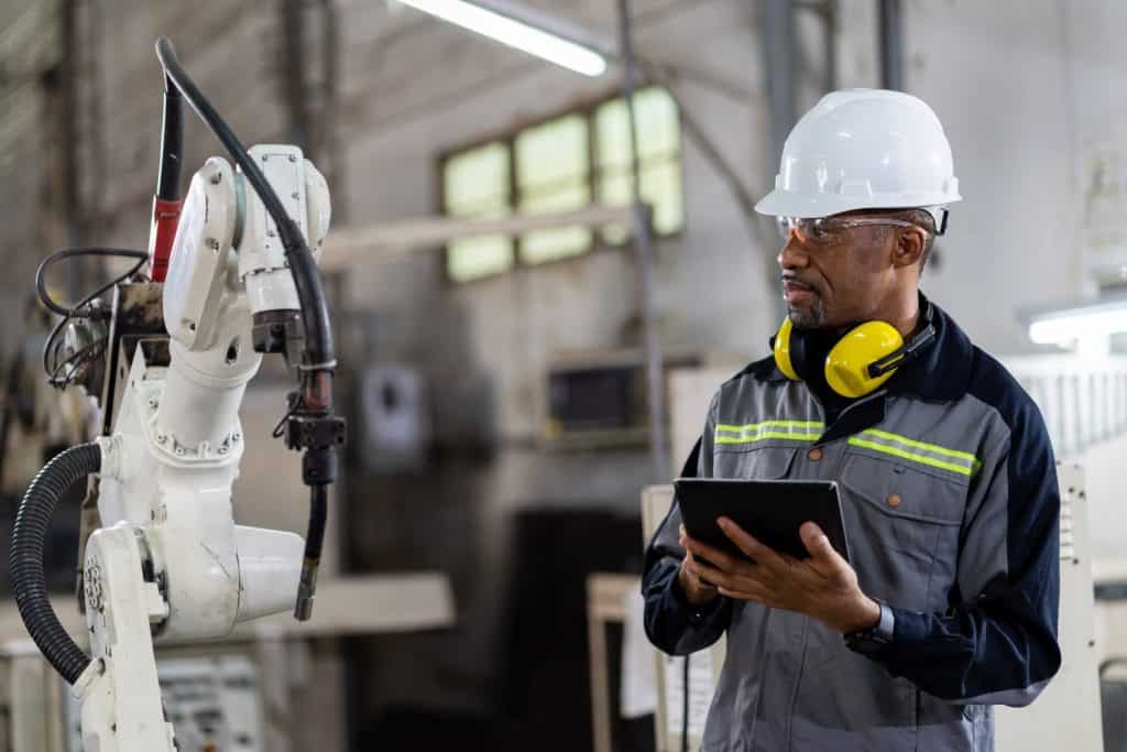 African American male engineer worker using digital tablet control automatic robotic hand machine in factory. Black male technician worker working with control automatic robot arm system welding