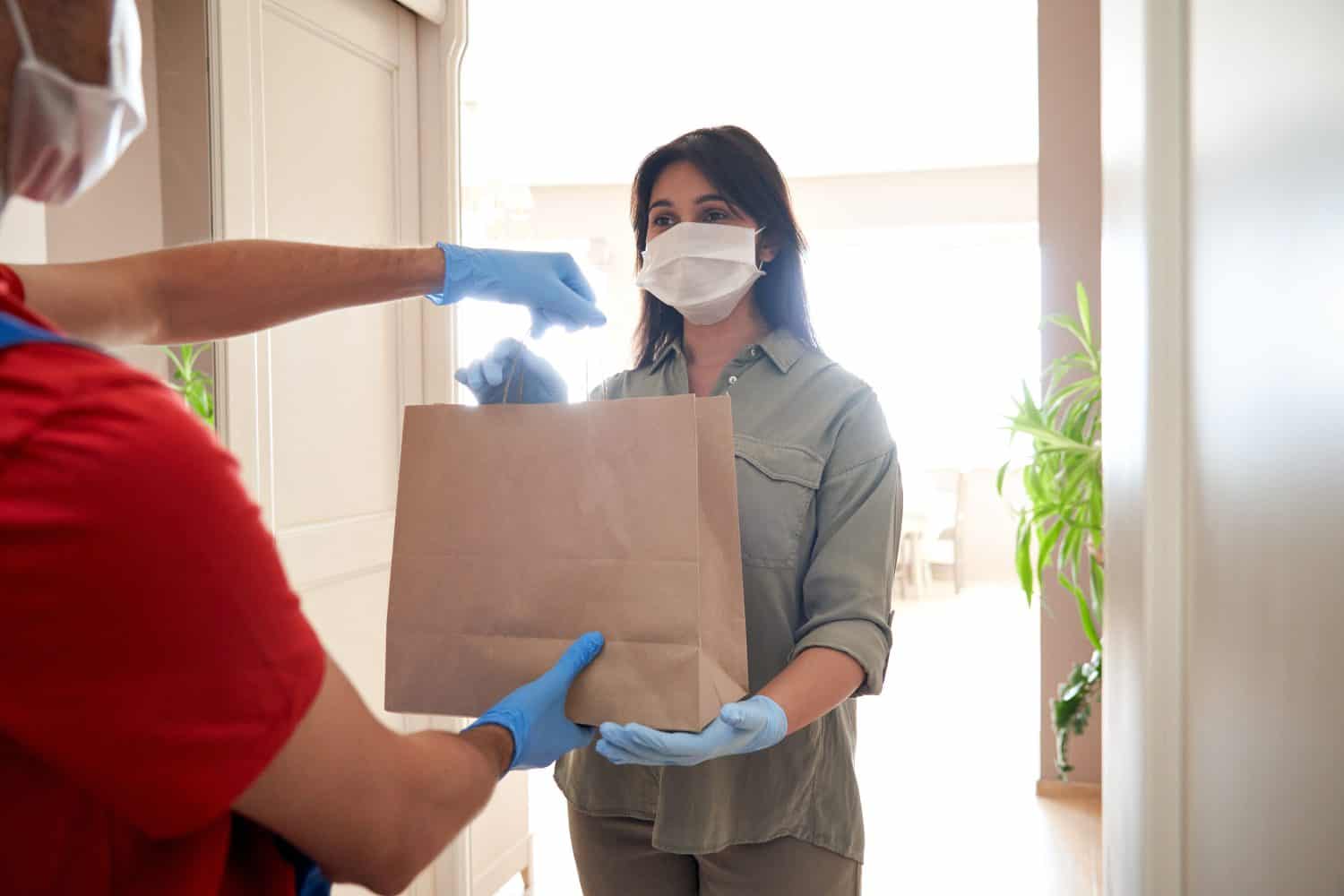 Indian woman customer wearing face mask and gloves taking delivery paper eco bag from man courier holding grocery food package delivering supermarket takeaway order standing at home. Safe delivery.