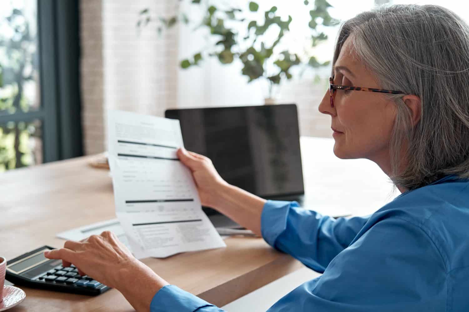 Senior mature business woman holding paper bill using calculator, old lady managing account finances, calculating money budget tax, planning banking loan debt pension payment sit at home office table.