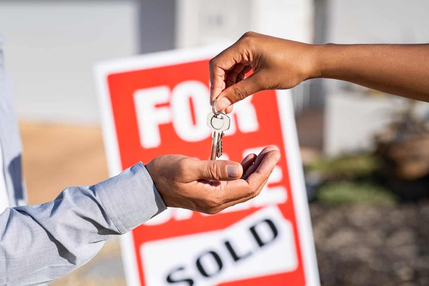 Saleswoman giving home keys to new property owner. Close up of real estate agent hand giving house keys to man. Agent giving apartment key to new owner after putting the word sold on for sale sign.