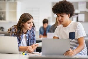 College students sitting together and using laptop during computer lesson. Black man and girl working on laptop in high school. Happy smiling multiethnic friends working on laptop for an assignment.