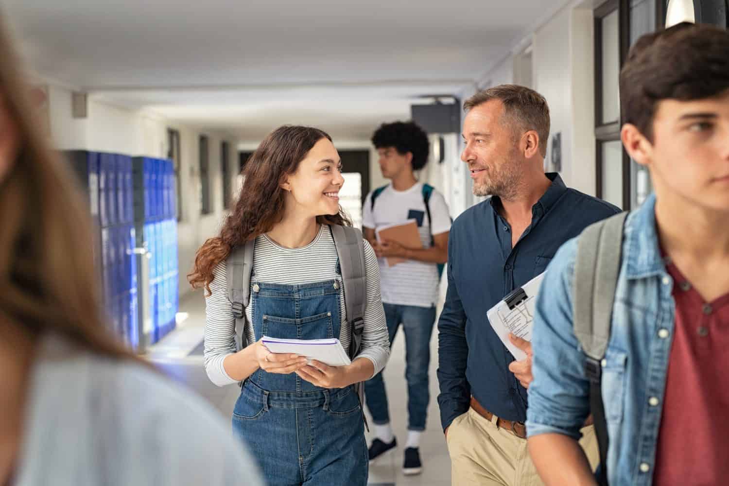 Happy high school student talking to mature man professor in hallway. Senior man teacher in conversation to school girl in campus. Helpful lecturer explaining concept to college student in corridor.