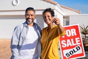 Portrait of happy mixed race couple holding house keys standing near sold signboard. Middle eastern man embracing african woman while showing house key outside of their new home. Moving home day.