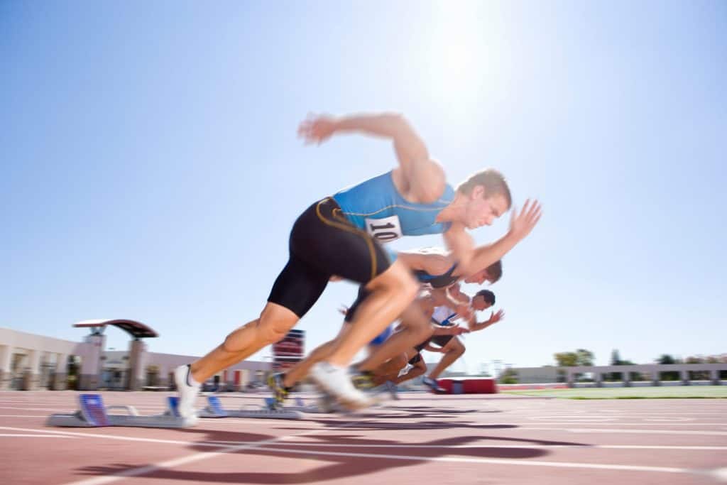 Side view of young male sprinters leaving their starting blocks at the start of a sprint race on a bright, sunny day at the track