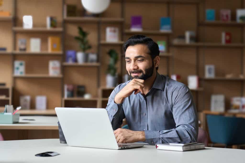 Smiling indian businessman working on laptop in modern office lobby space. Young indian student using computer remote studying, watching online webinar, zoom virtual training on video call meeting.