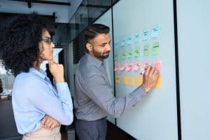 Young indian businessman manager writing strategy ideas on sticky notes on whiteboard and female African American colleague looking at his strategy scrum presentation. Business project planning.