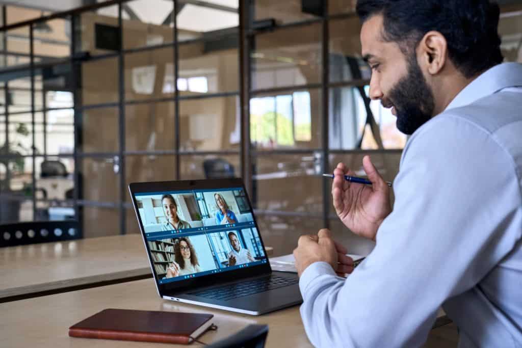 Indian happy smiling businessman ceo having working videocall on planning finance with multiracial team colleagues using laptop pc in modern corporation office. Over shoulder view.