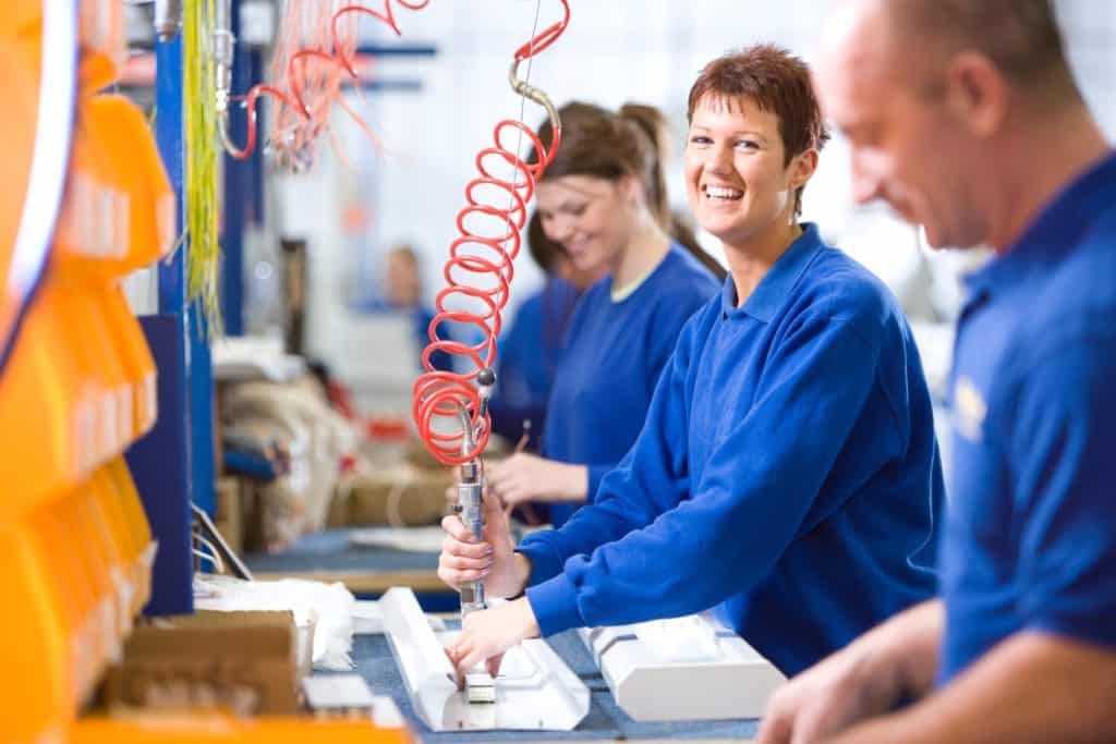 A medium shot of a young female worker smiling at camera while working on an aluminum light fittings on the production line with other workers.