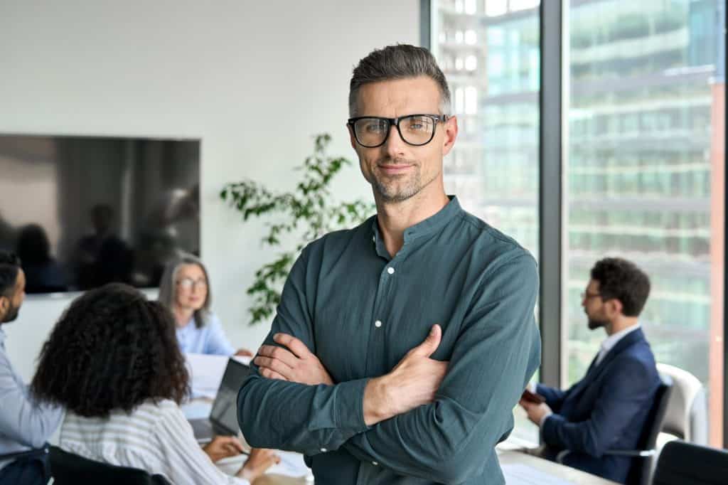 Smiling confident mature businessman leader looking at camera standing in office at team meeting. Male corporate leader ceo executive manager wearing glasses posing for business portrait arms folded.