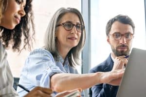 Mature older ceo businesswoman mentor in glasses negotiating growth business plan with diverse executive managers at boardroom meeting table using laptop. Multicultural team work together in office.