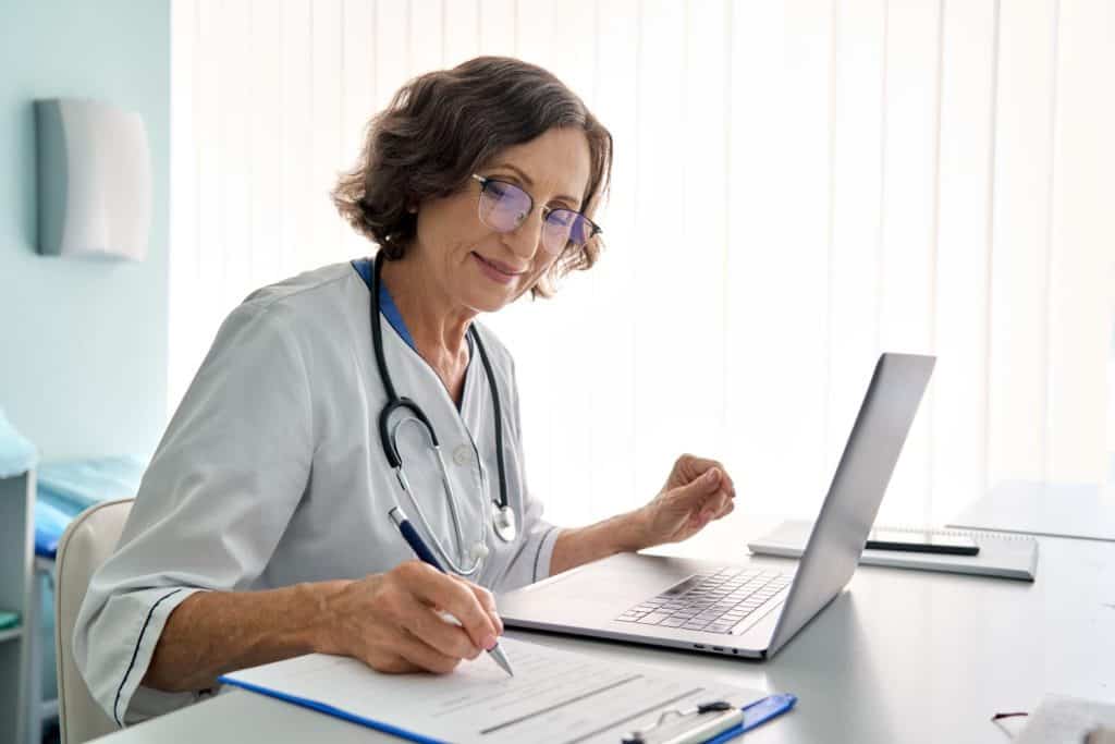 Senior middle aged female medical worker in modern clinic wearing white doctor's coat having videocall using laptop writing health personal data, consulting remotely. Telemedicine healthcare concept.