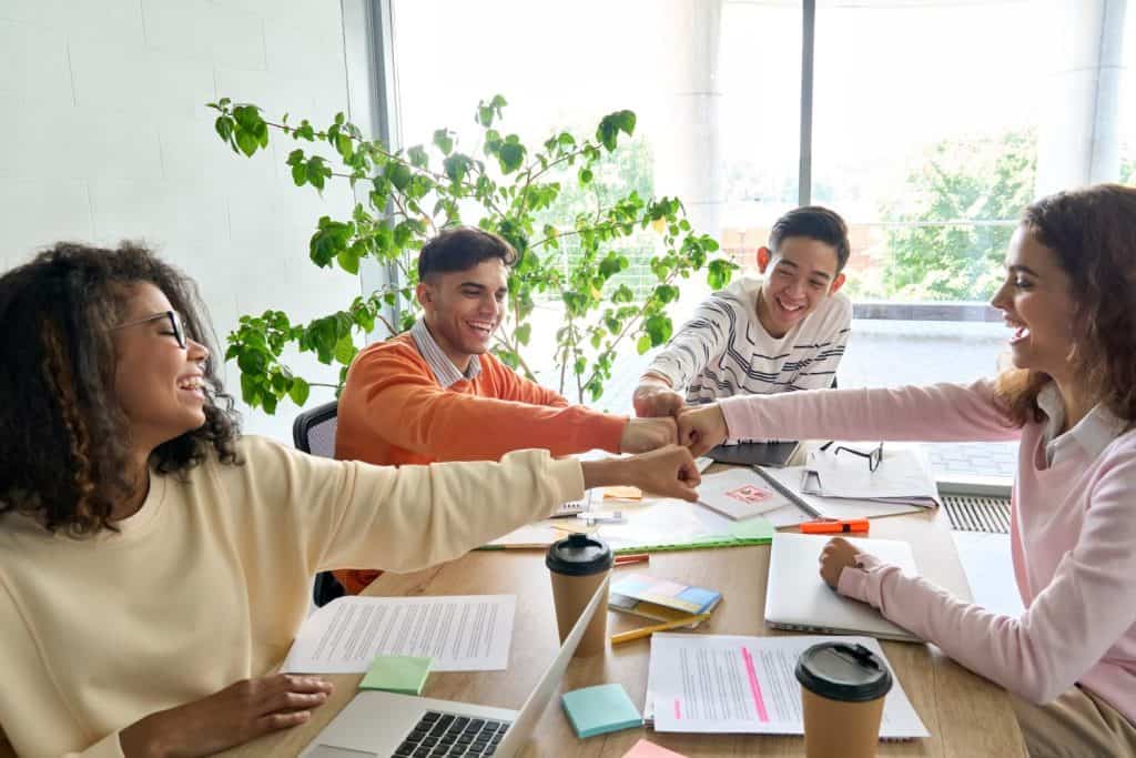 Young happy laughing creative startup team four multiethnic coworkers diverse students work together give fist bump celebrate successful project in office classroom at desk. Teamwork success concept.